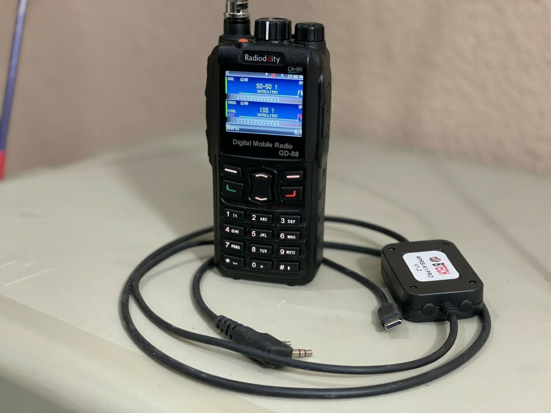 Auto-generated description: A digital mobile radio device with a connected charging or data transfer cable is resting on a surface.