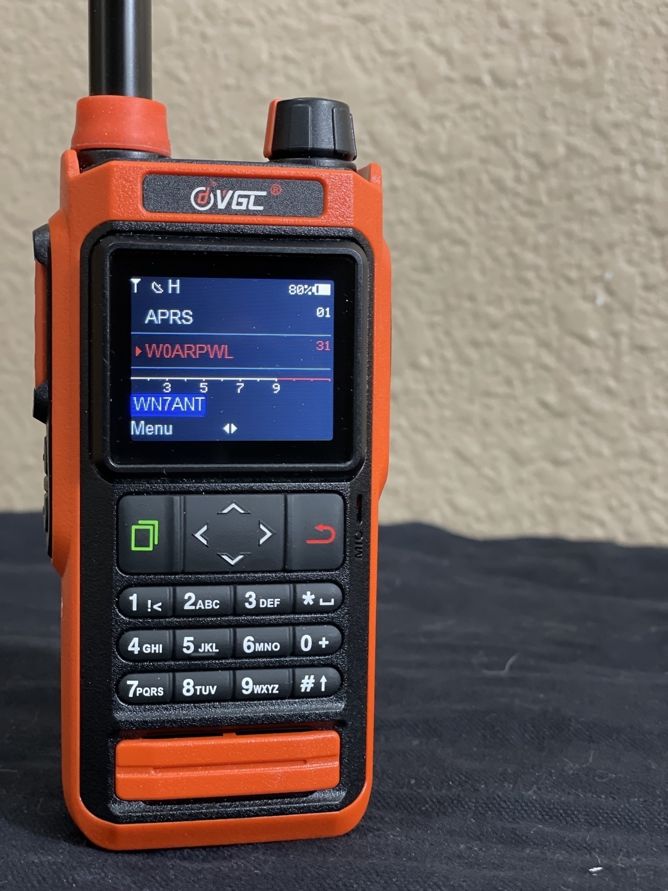 Auto-generated description: A handheld VGL radio device with a digital screen and numeric keypad is placed on a dark surface.